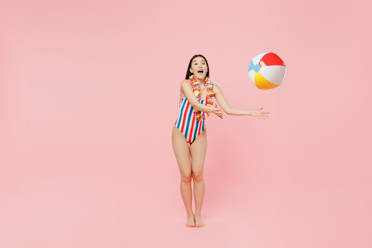 Full body young fun woman of Asian ethnicity in striped one-piece swimsuit hawaii lei toss up inflatable rubber ball isolated on plain pastel pink background. Summer vacation sea rest sun tan concept.