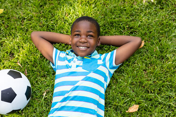 Smiling african american boy lying with hands behind head by soccer ball on grass in backyard