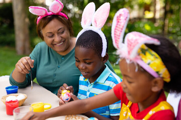 Obraz premium Happy african american siblings and grandpmother in bunny ears painting easter eggs in backyard