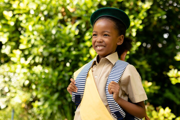 Smiling african american scout girl in uniform with backpack looking away against plants - obrazy, fototapety, plakaty