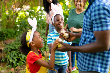 Naklejka premium Happy african american siblings in bunny ears giving easter eggs to father in backyard