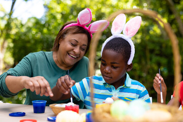 Fototapeta premium African american boy and grandmother wearing bunny ears while painting eggs on easter day