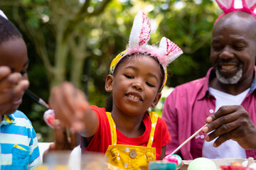 African american siblings and grandfather in bunny ears painting eggs on easter day