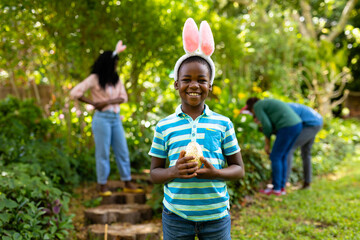 Fototapeta premium Portrait of happy african american boy in bunny ears holding easter egg while family in backyard