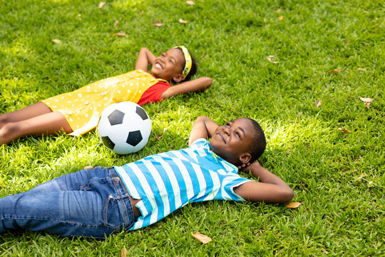 Smiling african american boy and girl lying with hands behind head by soccer ball in backyard