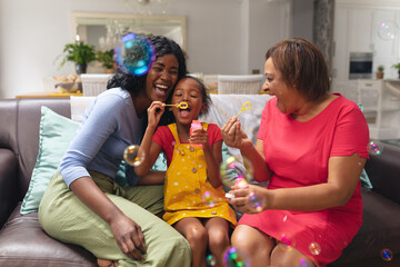 Playful african american girl blowing bubbles with mother and grandmother at home