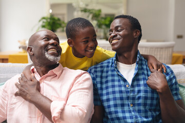 Happy african american boy with father and grandfather sitting on sofa at home - Powered by Adobe