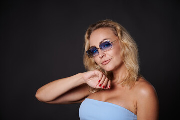 Confident portrait of a charming middle aged Caucasian woman with wavy blond hair wearing a blue top and blue sunglasses and looking at camera isolated over black background with copy ad space.