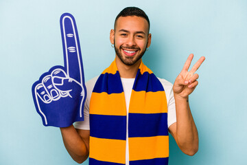 Young sports hispanic fan man isolated on blue background joyful and carefree showing a peace...