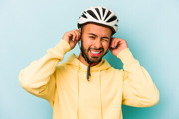Young hispanic man wearing helmet isolated on blue background covering ears with hands.