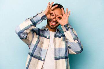 Young hispanic man isolated on blue background showing okay sign over eyes