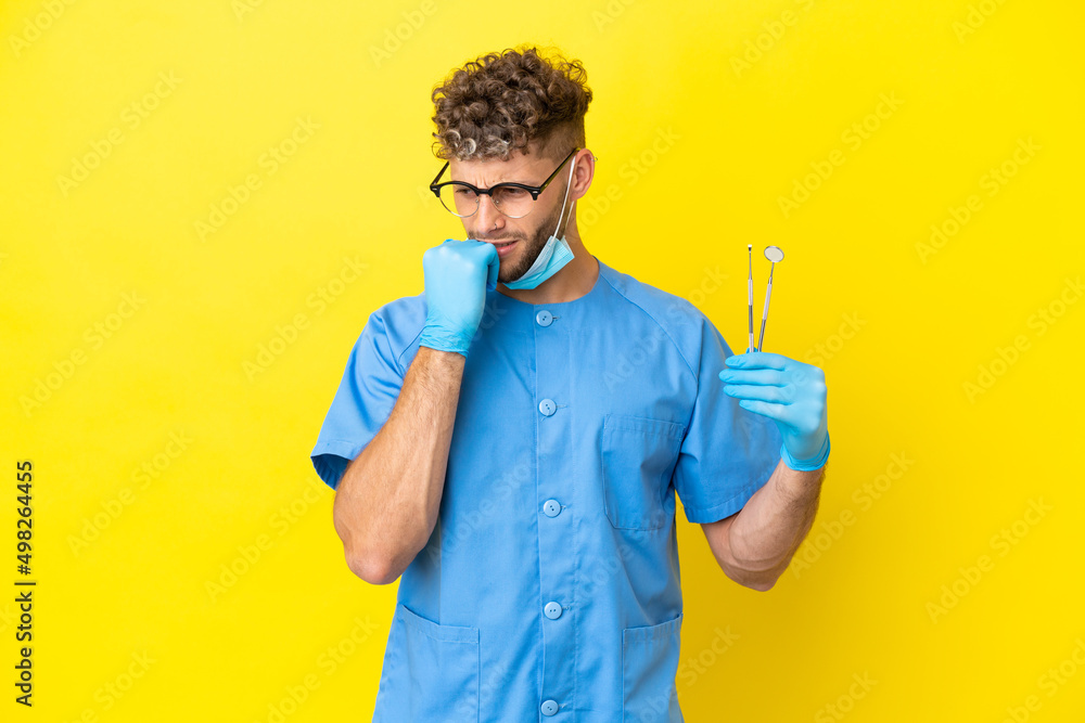 Wall mural Dentist blonde man holding tools isolated on background having doubts