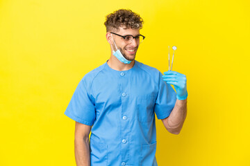 Dentist blonde man holding tools isolated on background looking to the side and smiling