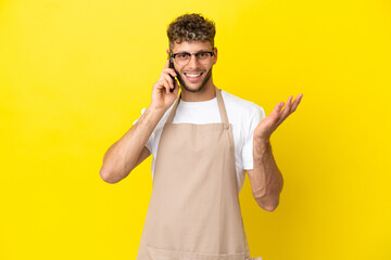 Restaurant waiter blonde man isolated on yellow background keeping a conversation with the mobile phone with someone