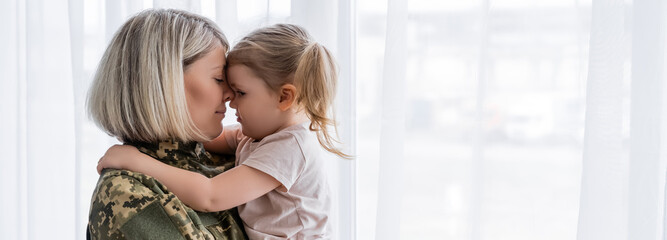 side view of military woman and girl embracing face to face near white window curtain, banner.