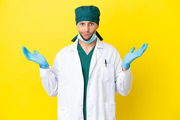 Surgeon blonde man in green uniform isolated on yellow background having doubts while raising hands