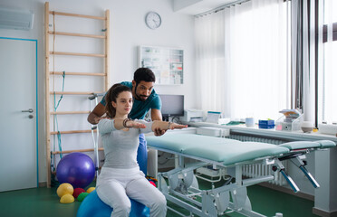 Young male physiotherapist exercising with young woman patient on ball in a physic room - obrazy, fototapety, plakaty