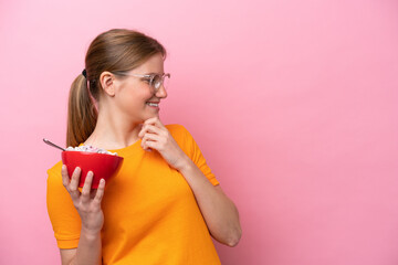 Young caucasian woman holding a bowl of cereals isolated on pink background thinking an idea and looking side