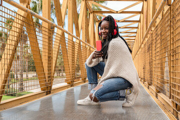 a young afro-american woman in braids listening to music on her cell phone with red headphones squatting on a yellow bridge