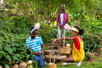 Fototapeta premium Happy african american siblings in bunny ears hiding easter eggs with grandfather in backyard