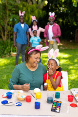 Happy african american girl and grandmother painting easter eggs while family in background