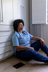 Stressful african american mid adult female nurse with eyes closed sitting on floor in hospital