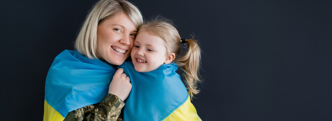 happy woman looking at camera near daughter and ukrainian flag isolated on black, banner.