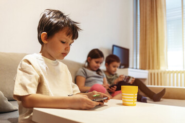 Group of kids using smartphone sitting on a sofa at home. Young boys and a girl sharing photo and video watching social story online. Friends enjoying new trend technology. Youth and tech concept.