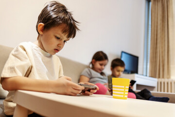 Three kids are sitting on couch and playing with modern mobile devices. Small group of children sit on sofa and use technology at home during the day. Group of kids using wireless technology at home.