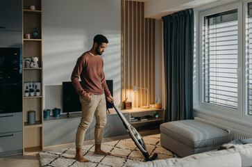 Young man hoovering carpet with vacuum cleaner in living room