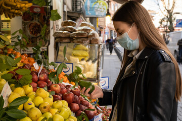 A young European girl with long dark hair buys fresh fruits and vegetables at the market in a protective mask to protect herself and others from covid-19. Woman is choosing avocado and pears.
