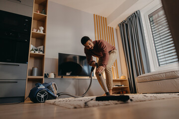 Young man hoovering carpet with vacuum cleaner in living room - obrazy, fototapety, plakaty