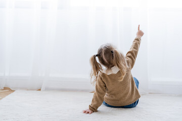 back view of little girl with ponytails pointing with finger while sitting on floor at home.