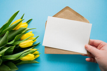 Hand holds envelope and paper for text on blue background and yellow tulips. Happy holiday greetings