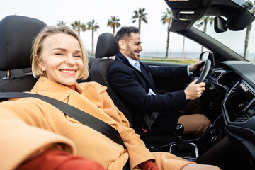 Smiling mid adult couple driving a car on road trip at the beach - Rental car service concept with...