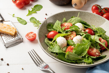 Fresh salad with arugula, cherry tomatoes, mozzarella cheese and hard cheese on white wooden background. Top view