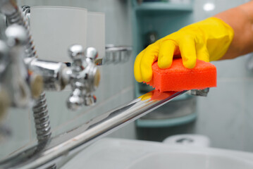 Bathroom washing, home cleaning. Close-up of hand in yellow rubber glove cleaning chrome faucet with sponge. Selective focus on foreground
