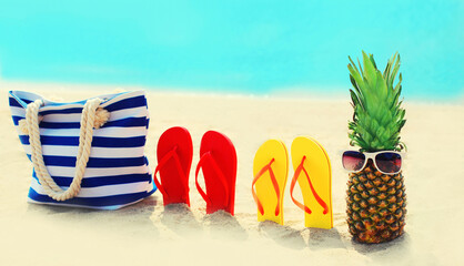Naklejka premium Summer vacation concept - bag with pineapple and flip flops on beach over blue sea background