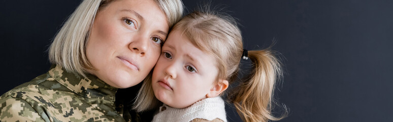 blonde military woman looking at camera near sad daughter isolated on black, banner.