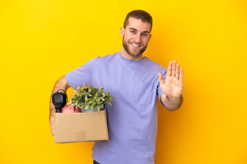 Young caucasian making a move while picking up a box full of things isolated on yellow background saluting with hand with happy expression