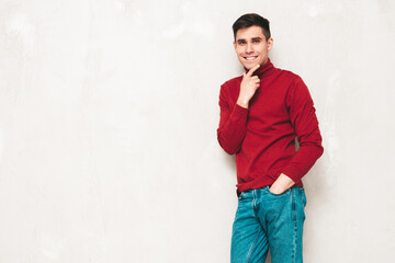 Fototapeta premium Portrait of handsome smiling model. Sexy stylish man dressed in red turtleneck sweater and jeans. Fashion hipster male posing near grey wall in studio. Isolated