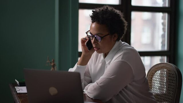 Female professional lawyer having mobile call and sitting at table with laptop in office spbas. 4k Closeup view of young african american woman talks on phone and looks at computer, discusses case and