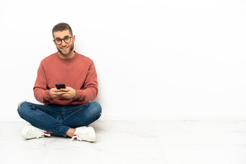 Young handsome man sitting on the floor sending a message with the mobile