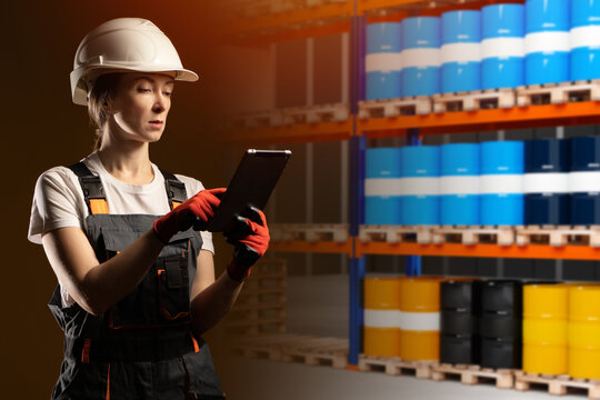 Oil warehouse. Woman manager of chemical products warehouse. Girl in work uniform with tablet. Storage and logistics of petrolium. Selective focus. Racks with oil drums of different colors