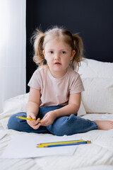 little girl sitting on bed near blue and yellow pencils and looking at camera.