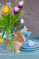 Little red decorative rabbit on a table decorated for Easter with eggs and a bouquet of tulips.