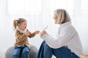 cheerful blonde woman holding card near amazed daughter while sitting on poufs at home.