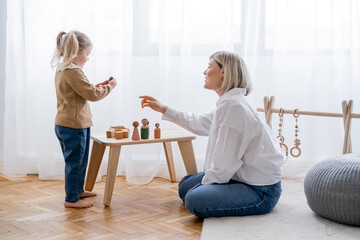 side view of woman pointing with hand near daughter playing with wooden figurines at home.
