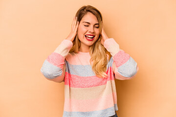 Young caucasian woman isolated on beige background covering ears with hands trying not to hear too loud sound.