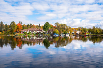 View of lakeside brick row houses surrounded by deciduous trees at the peak of fall foliage on a partly cloudy morning. Reflection in water.